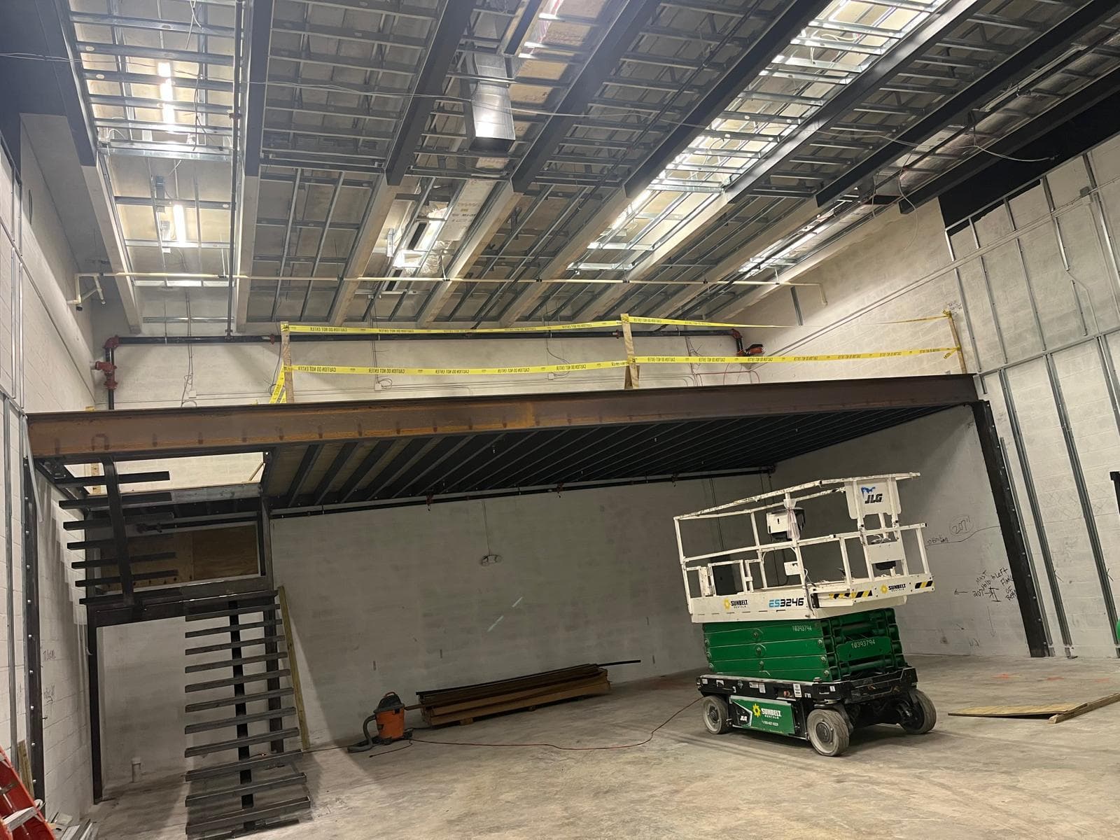 Warehouse interior during construction of a steel mezzanine and stair, with a scissor lift on the concrete floor