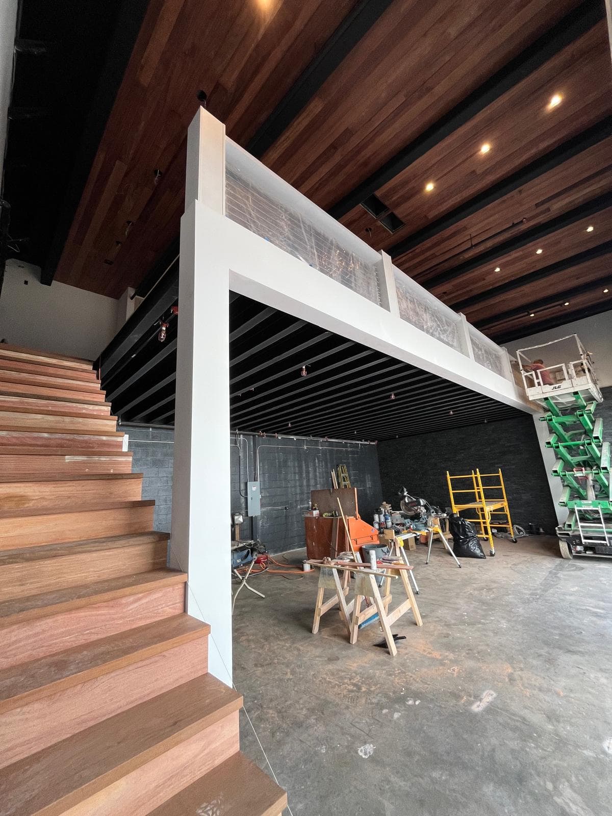 Wood-tread staircase and white landing beam beneath a stained wood-plank ceiling with black steel beams